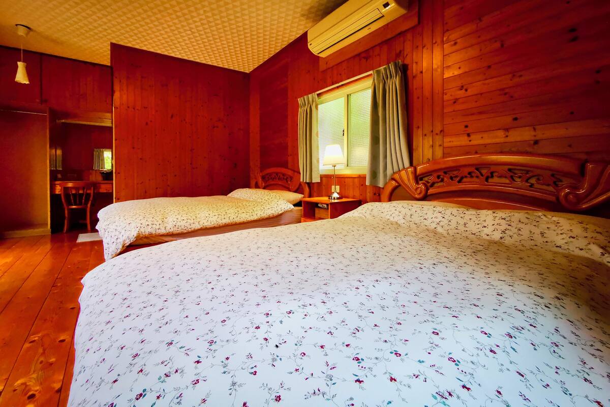 A room with two beds, wooden walls, a desk, and a window with curtains.