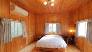 A wooden-paneled bedroom with a bed, two windows with curtains, a desk with a lamp, and an air conditioning unit. - Fong Chou Cottage (Sanyi)