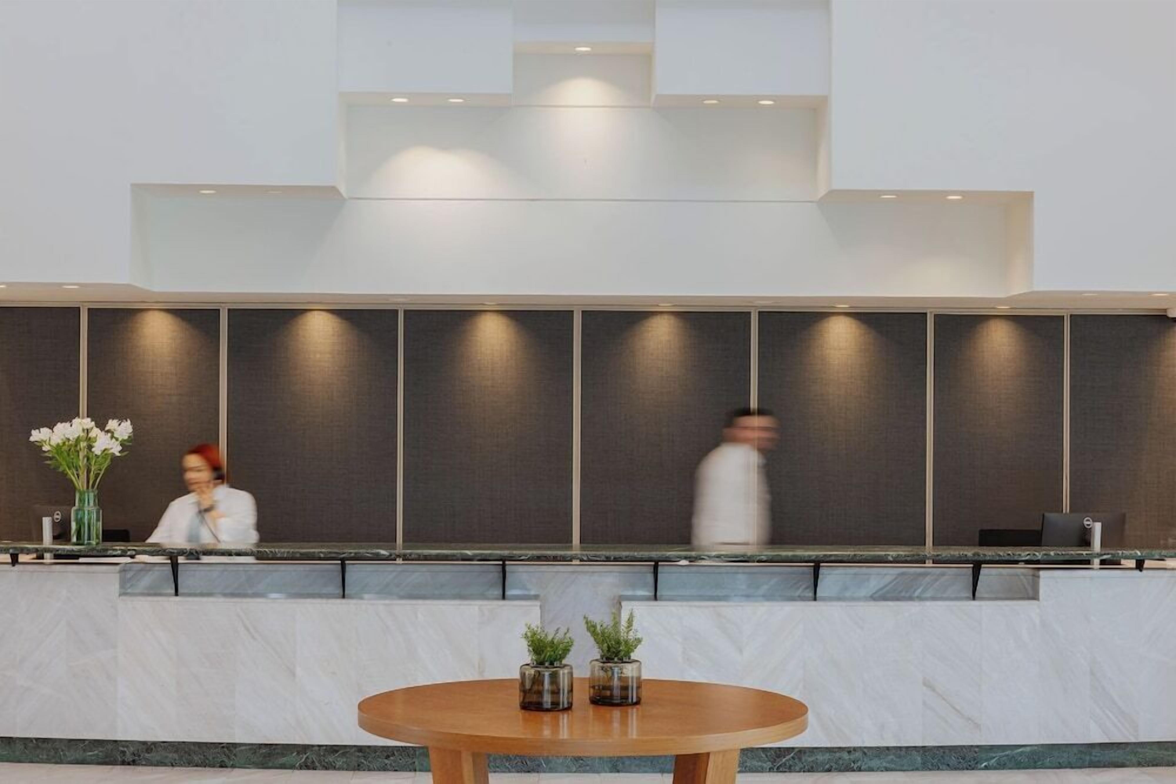 Una moderna zona de recepción con una barra de mármol, una mesa redonda de madera con plantas en macetas y una persona al fondo.