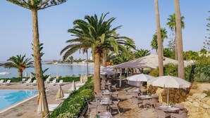 A poolside area with lounge chairs and umbrellas, a stone wall, and a view of the sea. - Nissi Beach Resort (Ayia Napa)