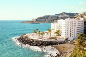 A coastal hotel with a rocky shoreline, palm trees, and a view of the sea and distant buildings. - Sunwing Arguineguin (Mogan)