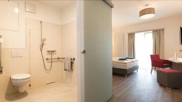 A modern hotel room with a toilet, shower, bed, and a small sitting area with a chair and table.