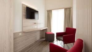 Una habitación de hotel moderna con un televisor de pantalla plana montado en una pared de madera, un pequeño taburete gris y dos sillas rojas alrededor de una mesa de vidrio.