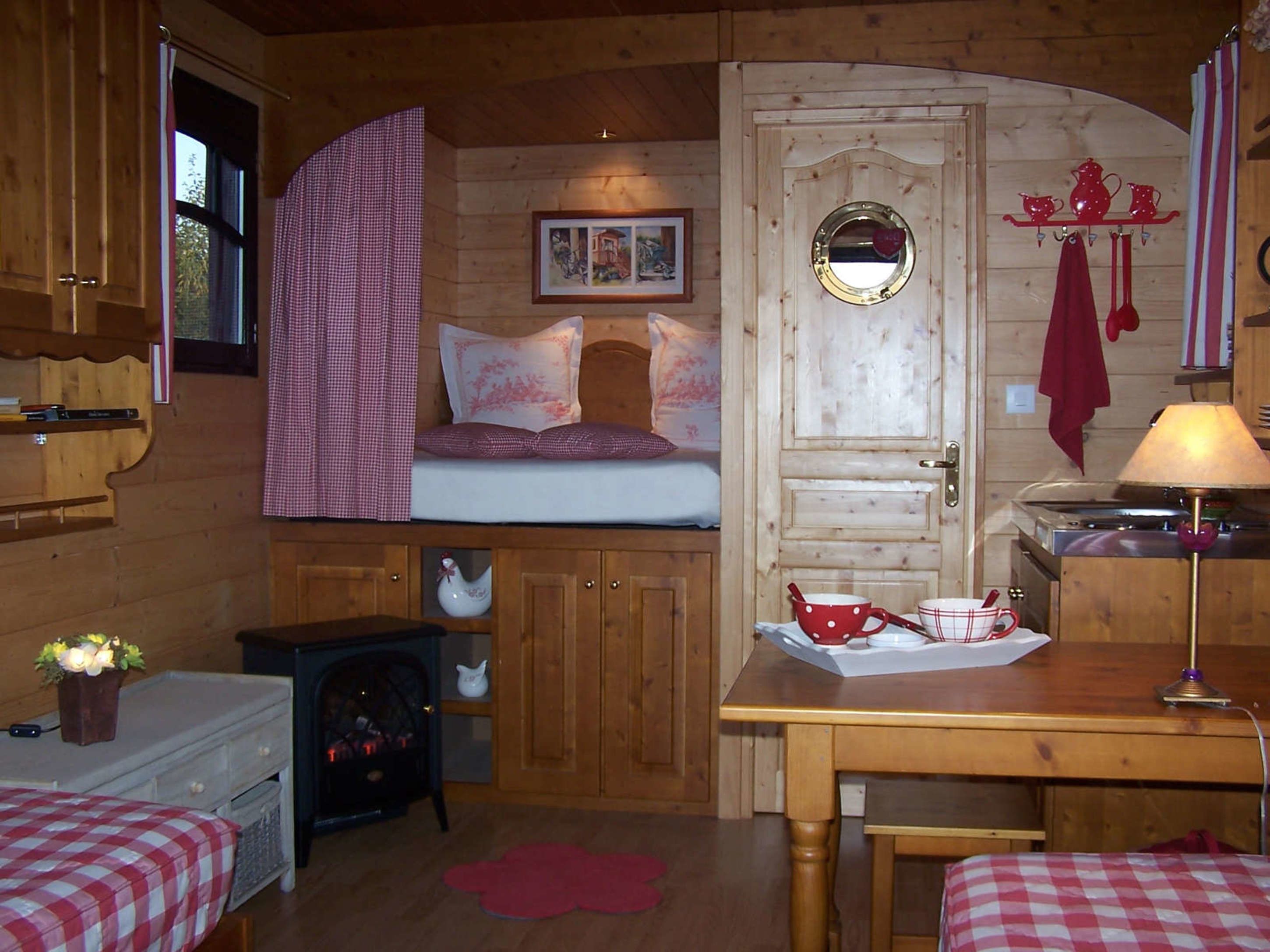 A cozy cabin-style room with a bed, a small table with a lamp, a fireplace, and a window with curtains.
