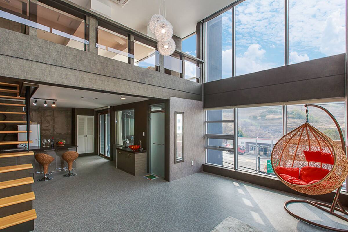 Un interior espacioso con techos altos, una escalera moderna, un ventanal grande con vistas al exterior y un sillón colgante con cojines rojos.