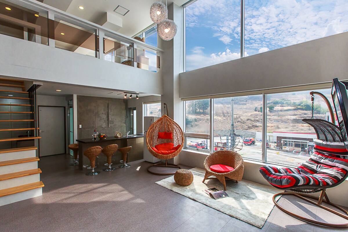 Un interior espacioso y moderno con techo alto, un entrepiso y ventanas grandes que ofrecen vistas del paisaje.