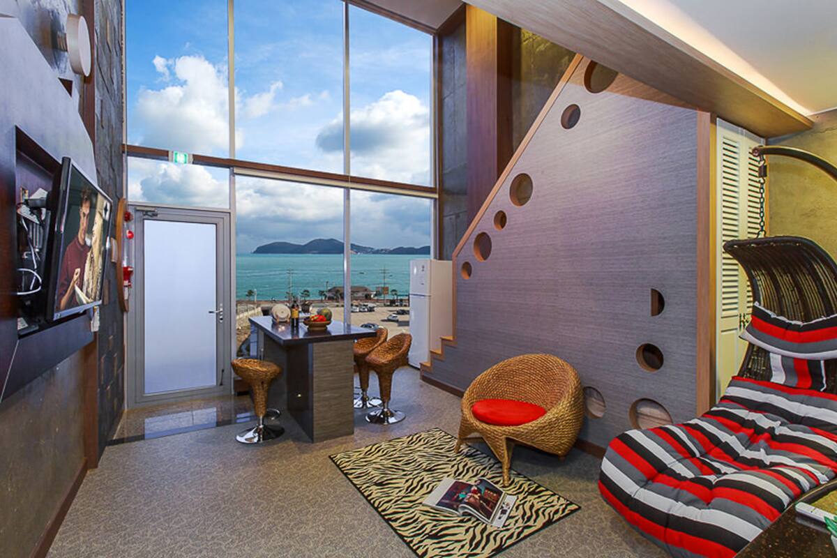Una habitación moderna con un ventanal grande que da a una playa, una zona de bar y una sala de estar con un cojín a rayas.