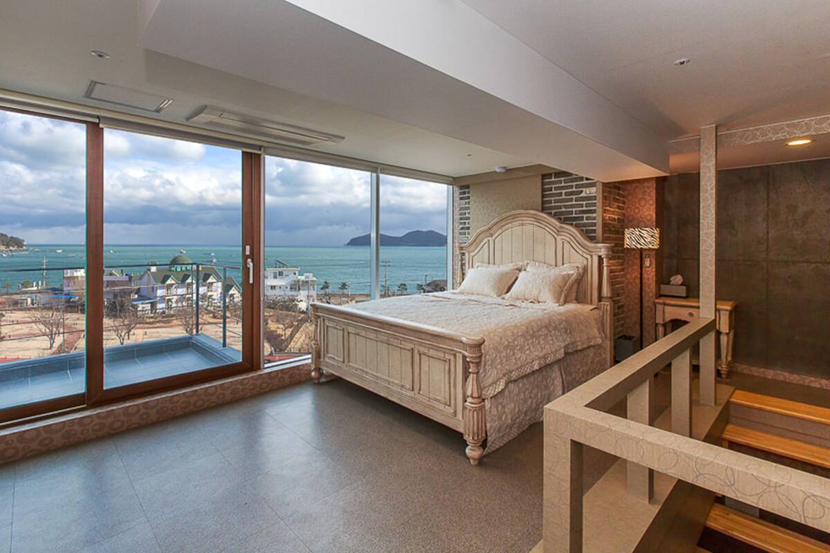Un dormitorio con un ventanal grande que da a una playa y al océano, una cama con cabecera apoyada en una pared de ladrillo, y un balcón con vistas al mar.