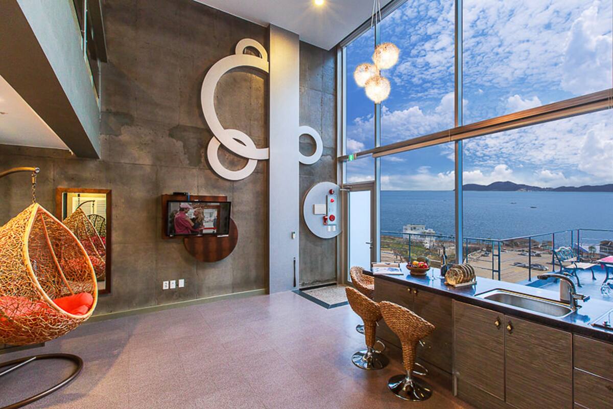 Una habitación moderna con un amplio ventanal que ofrece vistas panorámicas, un sillón colgante y una zona de bar con taburetes.