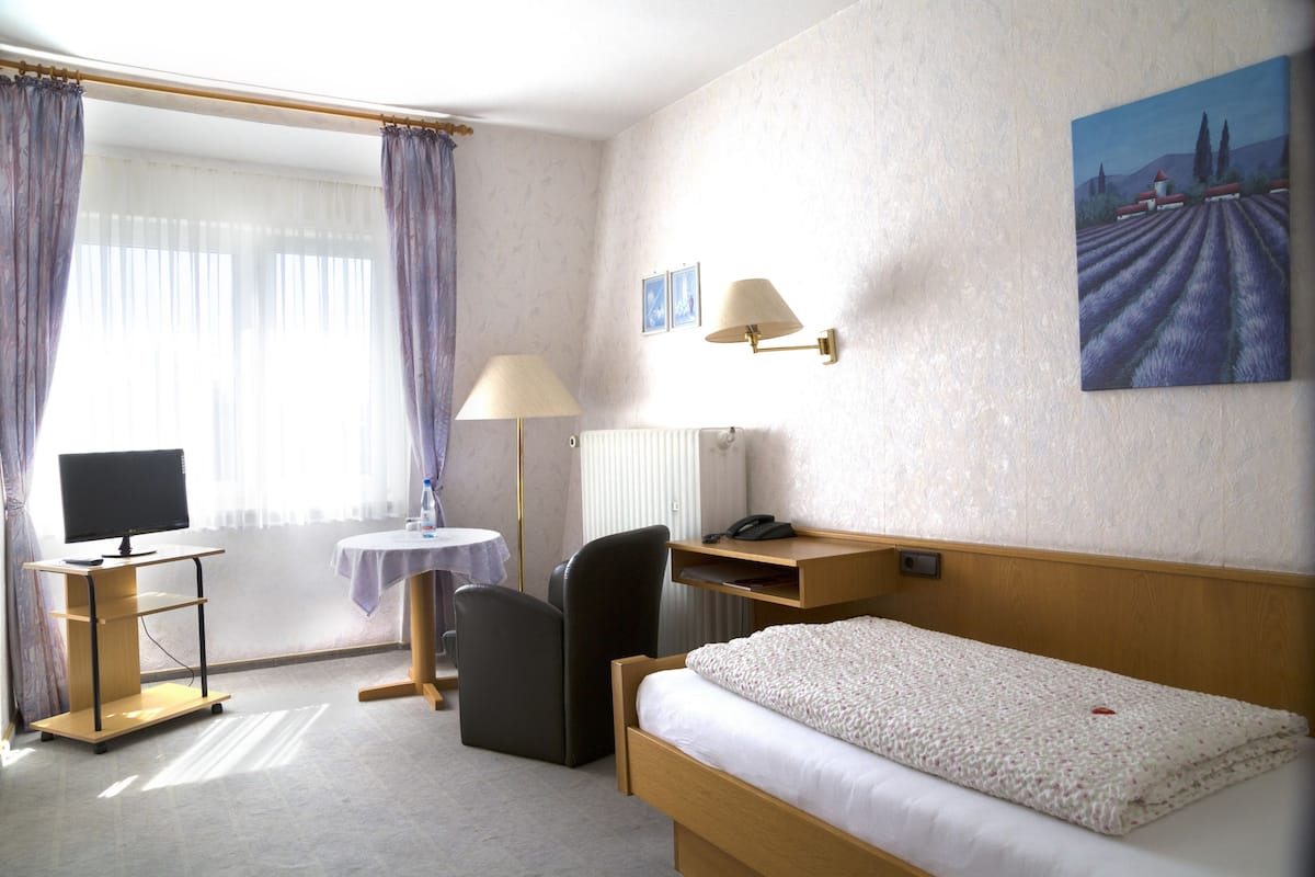 A hotel room with a bed, desk, chair, TV, lamp, and artwork on the wall.