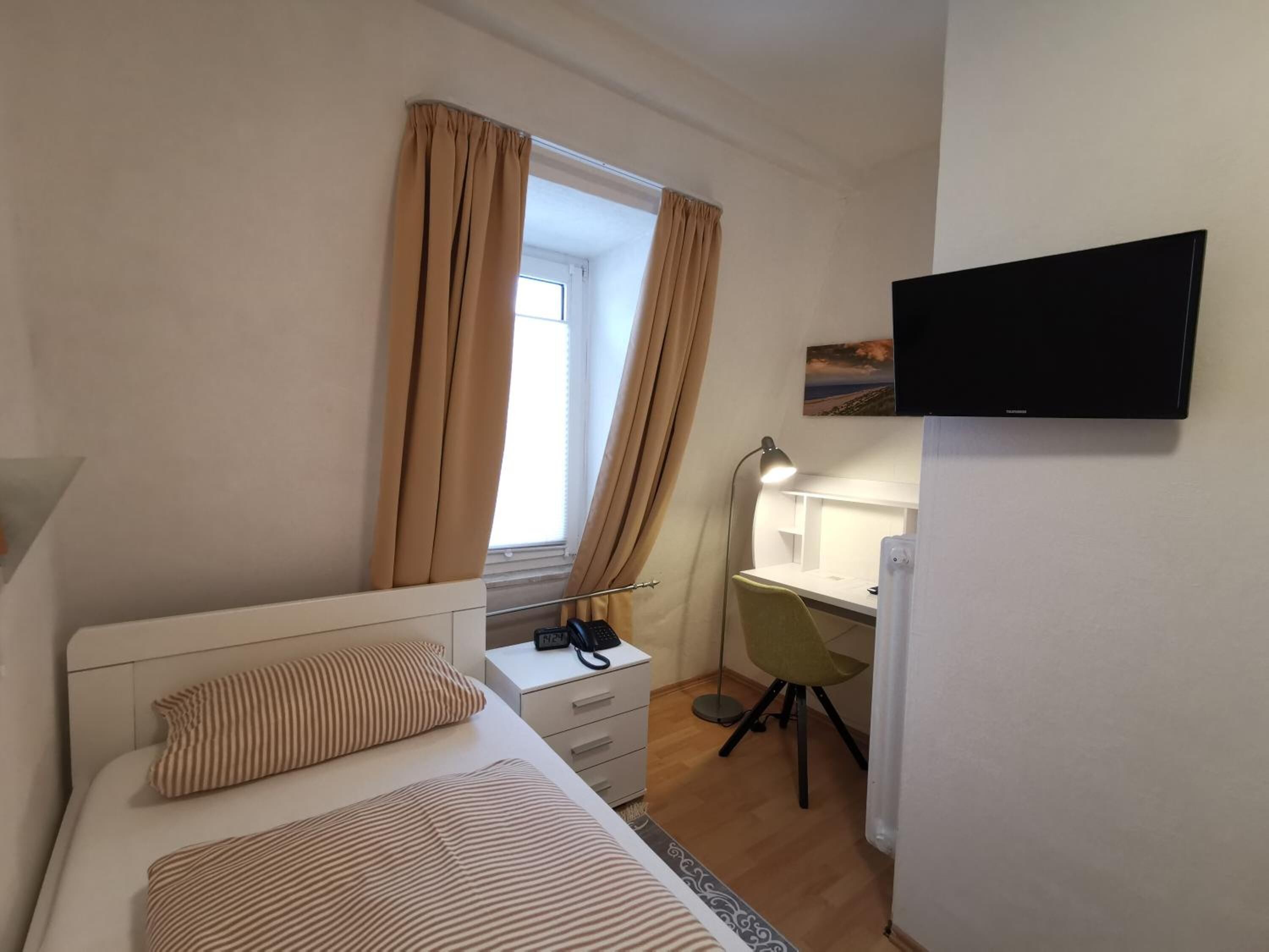 Ein kleiner, gut beleuchteter Raum mit einem Bett, einem Schreibtisch, einem Stuhl, einem Fernseher und einem Fenster mit Vorhängen.