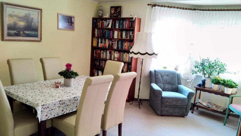 Un comedor con una mesa dispuesta para cuatro personas, rodeada de sillas. En el fondo, una estantería llena de libros, y una lámpara sobre una mesita auxiliar.