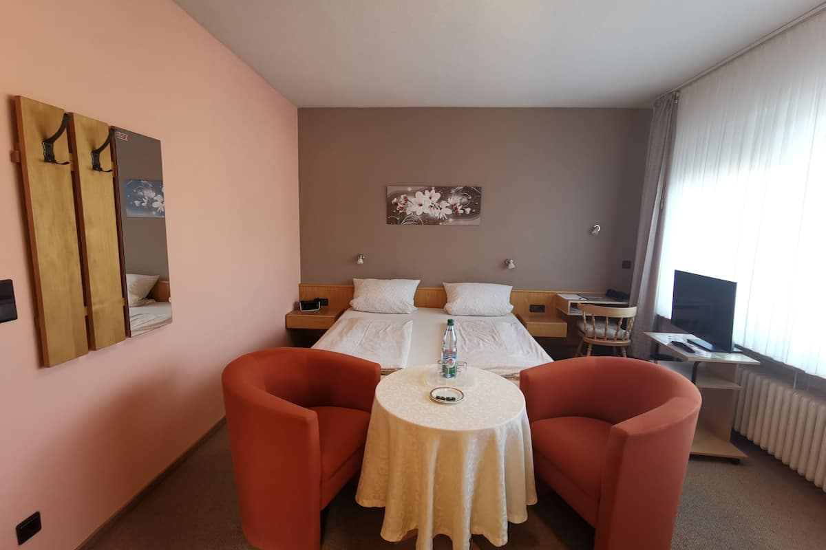 A hotel room with a bed, a desk with a TV, a table with chairs, a mirror, and a painting on the wall.