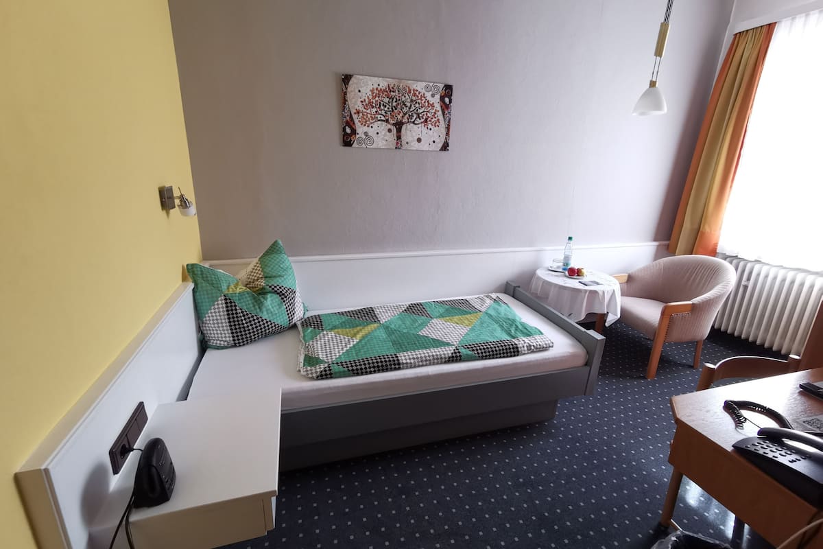 A compact room with a bed, a small table, a chair, a desk, a wall-mounted artwork, and a hanging light fixture.
