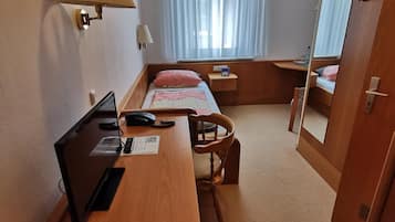 Ein kleines Hotelzimmer mit einem Holztisch, einem Stuhl, einem Bett mit rosa Kissen, einem kleinen Nachttisch, einer Lampe, einem Fenster mit durchscheinenden Vorhängen und einem Spiegel an der Tür.