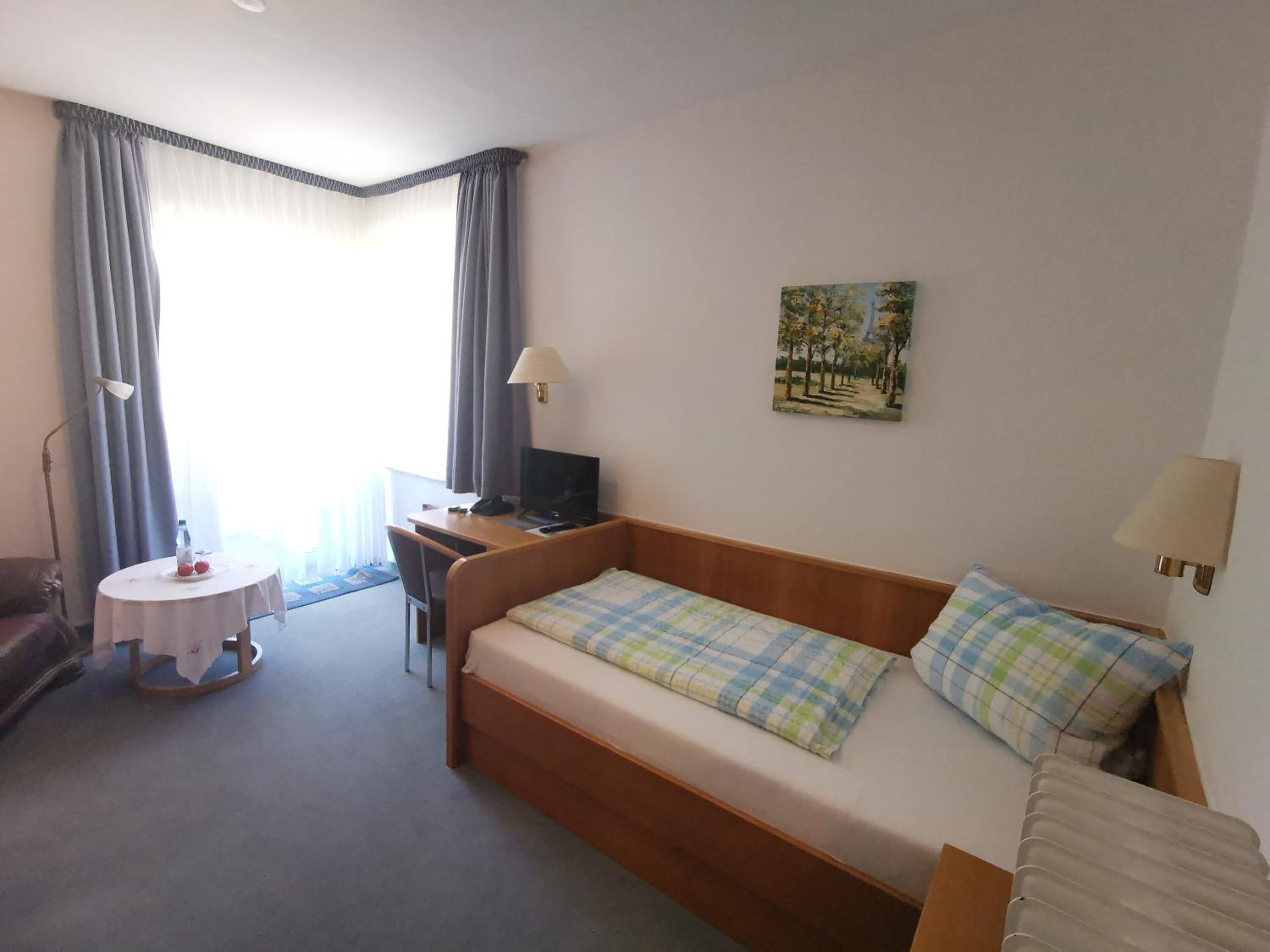 A hotel room with a bed, a desk, a chair, a small table with a bottle and glasses, and a painting on the wall.