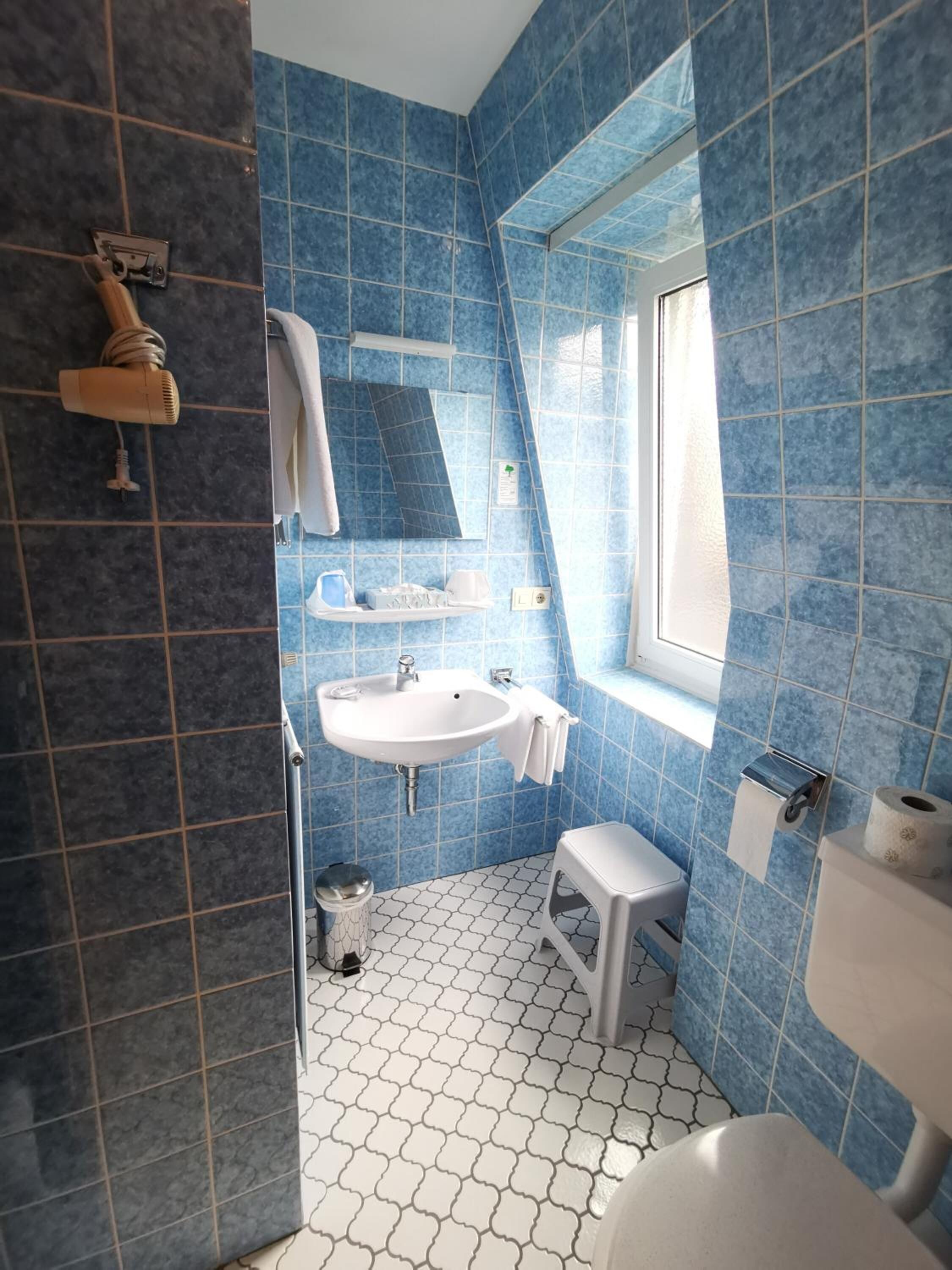 Ein Badezimmer mit blauen Fliesen, einem weißen Waschbecken, einem Spiegel, einem Fenster und einer Toilette.