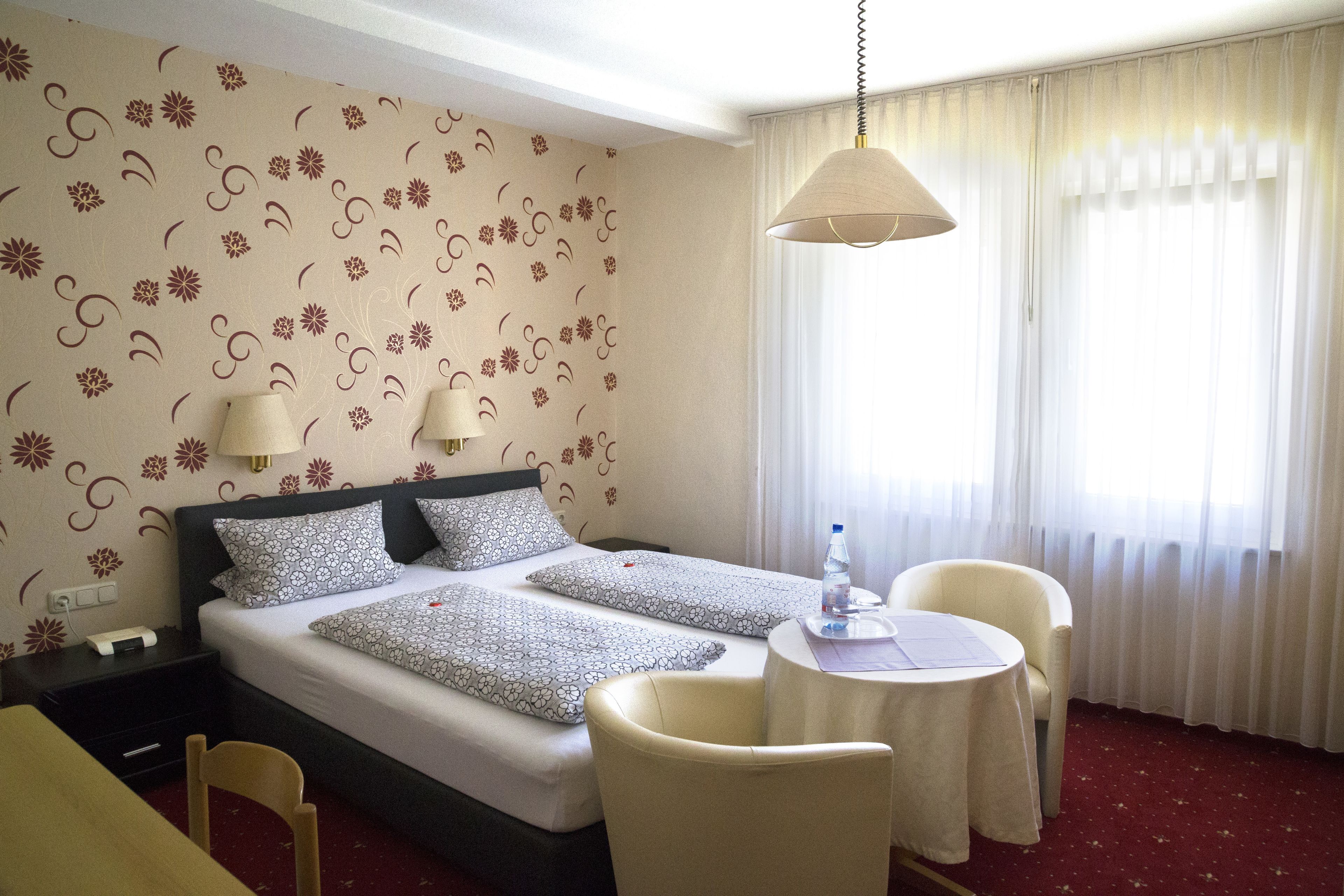 A hotel room with a double bed, two bedside tables with lamps, a small round table with a chair, and a window with sheer curtains.