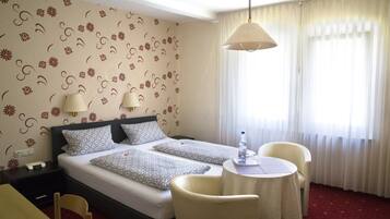 A hotel room with a double bed, two bedside tables with lamps, a small round table with a chair, and a window with sheer curtains.