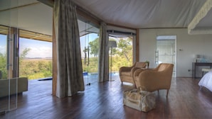 A spacious room with a large bed, a desk, and a seating area with chairs. There is a sliding glass door leading to an outdoor area with a view of the landscape. - Ngerende in the Wild Lakeview (Maasai Mara)