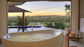 A bathroom with a large bathtub filled with bubbles, a view of a natural landscape, and a wooden vanity with a candle and glasses. - Ngerende in the Wild Lakeview (Maasai Mara)
