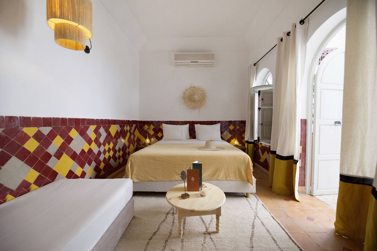 A hotel room with a bed, a small round table with a book and a hat, a wall air conditioner, and a patterned wall.
