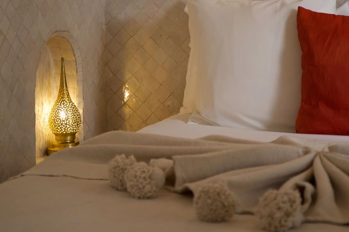 A neatly made bed with white and red pillows, a decorative wall with a patterned tile design, and a wall-mounted lamp.