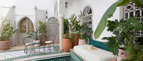 A courtyard with a pool, potted plants, a seating area, and a white building with arched windows and intricate metalwork.