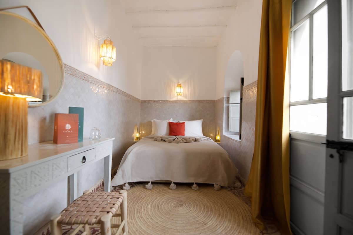 A bedroom with a bed, bedside lamps, a mirror, a bedside table, and a window.