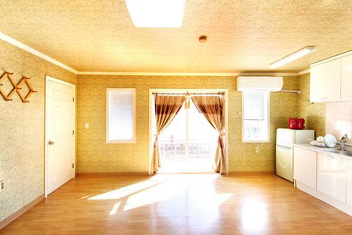 A spacious room with a wooden floor, a large window with curtains, a door, and a kitchenette area with a refrigerator and cabinets.