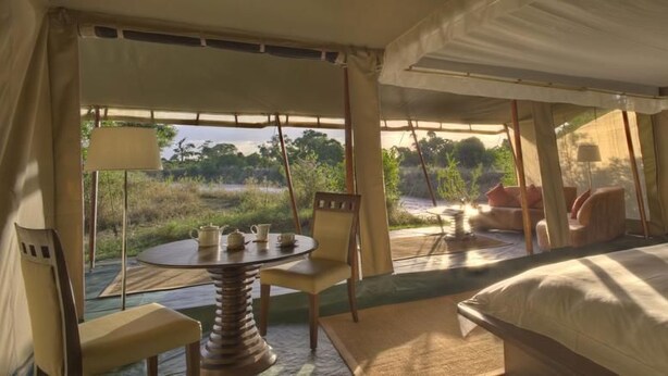 A tented room with a bed, a table with chairs, and a view of the outdoors. - Ngerende in the Wild Riverview (Maasai Mara)