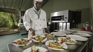 A chef preparing salads in a kitchen with stainless steel appliances and a tented outdoor area. - Ngerende in the Wild Riverview (Maasai Mara)