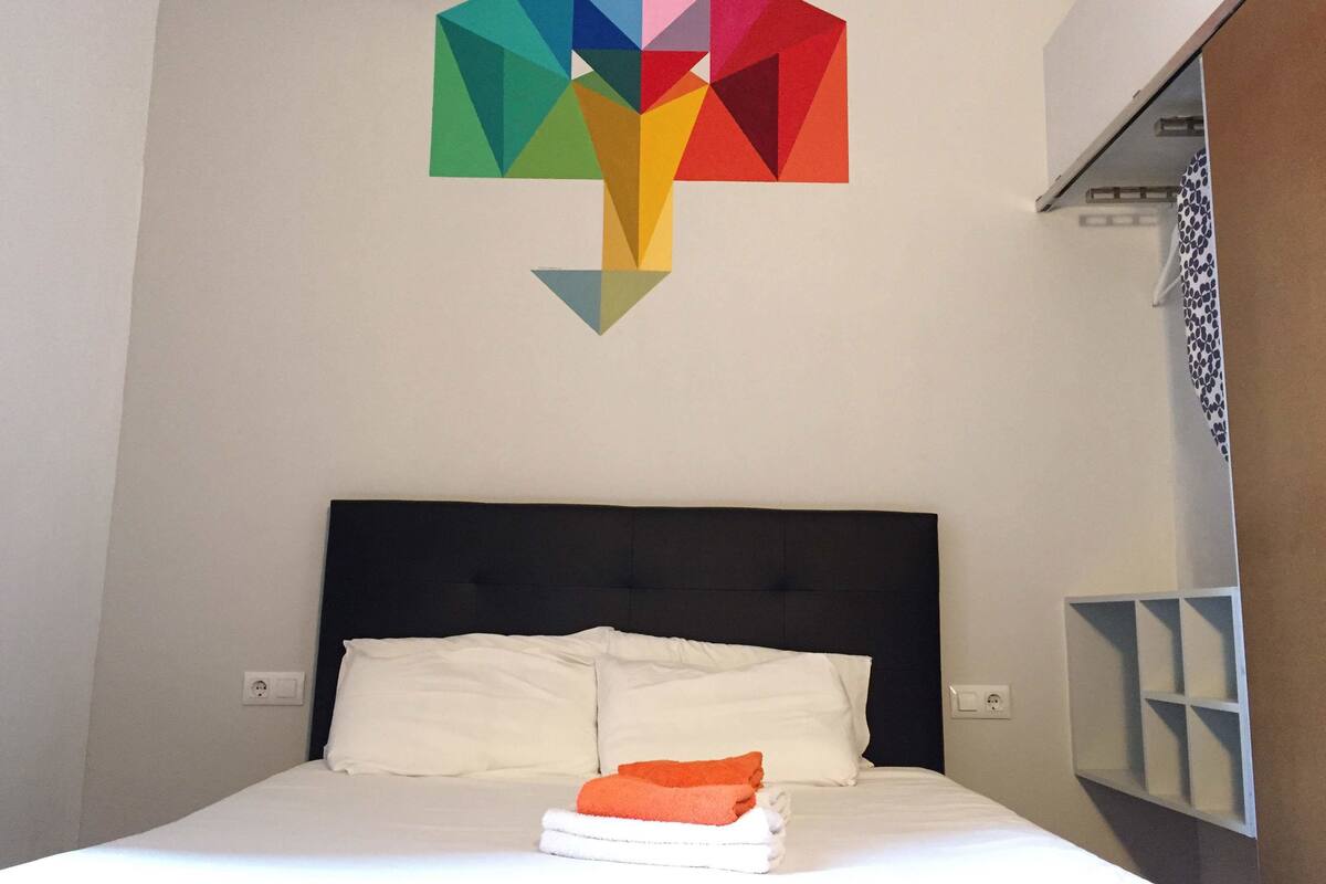 Een opgemaakt bed met witte lakens en twee kussens, een kleurrijk geometrisch kunstwerk erboven, en een patroon gordijn gedeeltelijk zichtbaar aan de rechterkant.