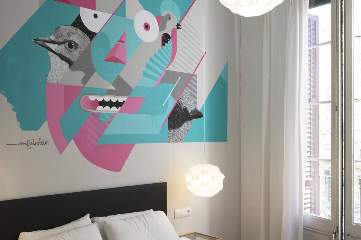 Een moderne hotelkamer met een groot abstract schilderij aan de muur, een net opgemaakt bed met witte lakens en twee gevouwen handdoeken, een bedlamp en uitzicht op de straat door een open deur.