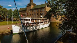 A white riverboat docked at a pier with a large building featuring a green dome in the background. - Hotel Slottsgården (Vadstena)