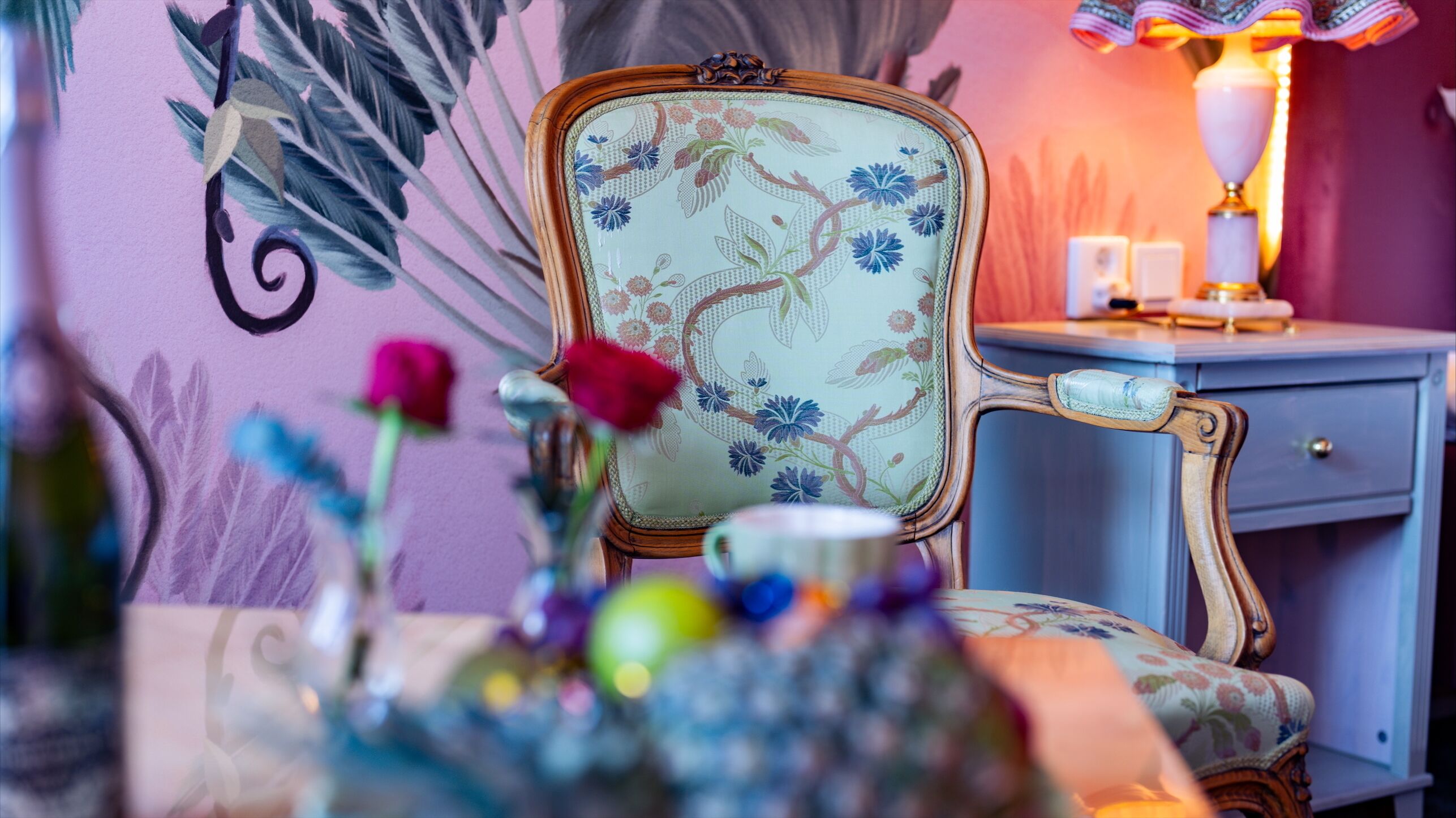 A room with a floral-patterned chair, a bedside table with a lamp, and a vase with flowers.
