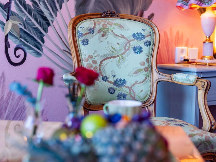A room with a floral-patterned chair, a bedside table with a lamp, and a vase with flowers.