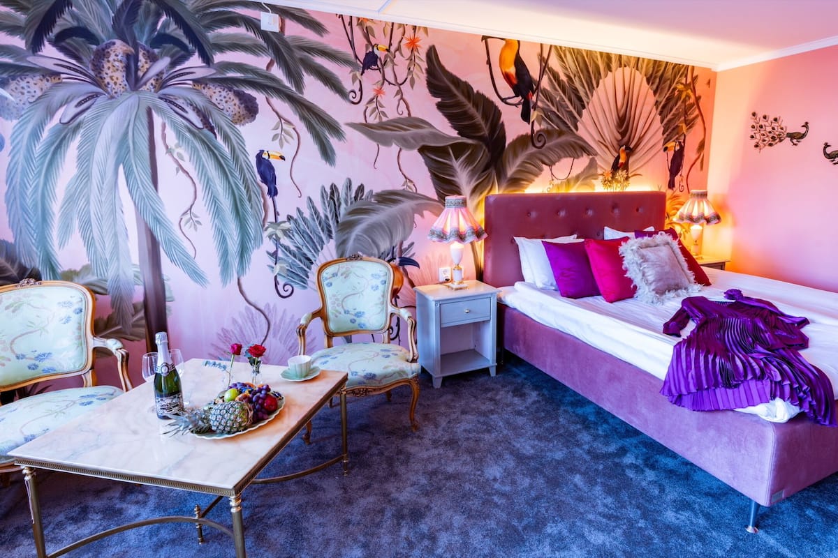 Ein Zimmer mit einem Bett, einem Nachttisch, einem Sessel, einem Tisch mit Obst und einem Wandbild von Vögeln und Blättern.