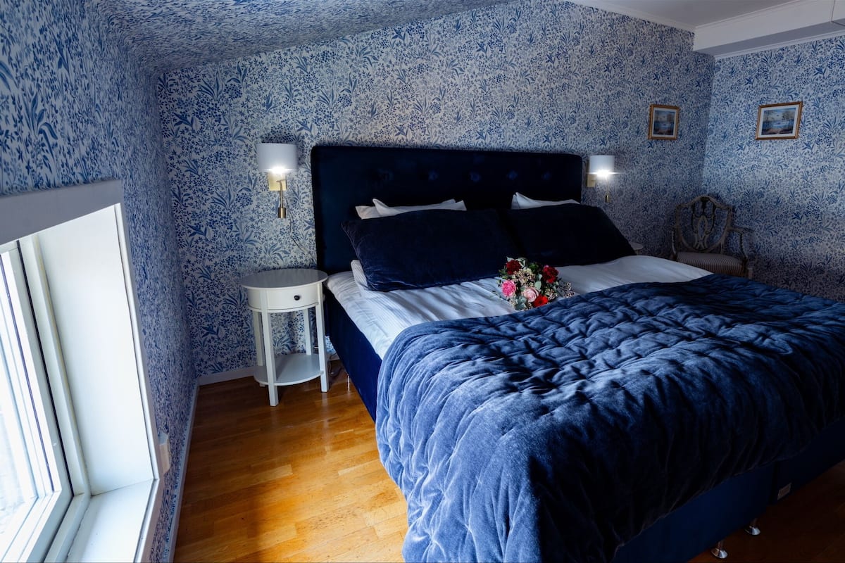 Ein Schlafzimmer mit floralem Wandpapier, einem dunkelblauen Bett mit weißer Bettwäsche, einem Nachttisch mit Lampe und einem kleinen Holzsessel.