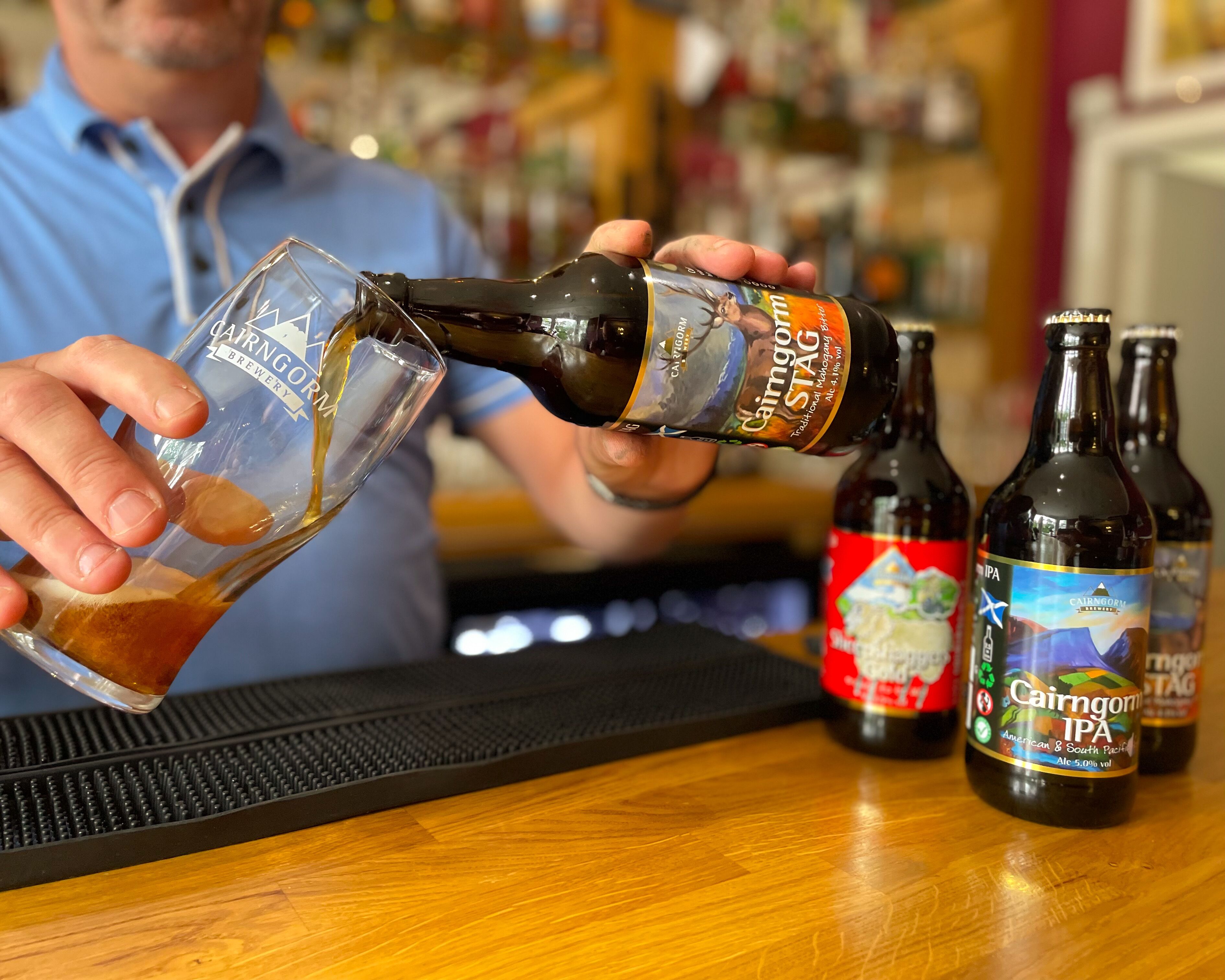 A person pouring a dark liquid from a bottle labeled "Cairngorm IPA" into a clear glass.