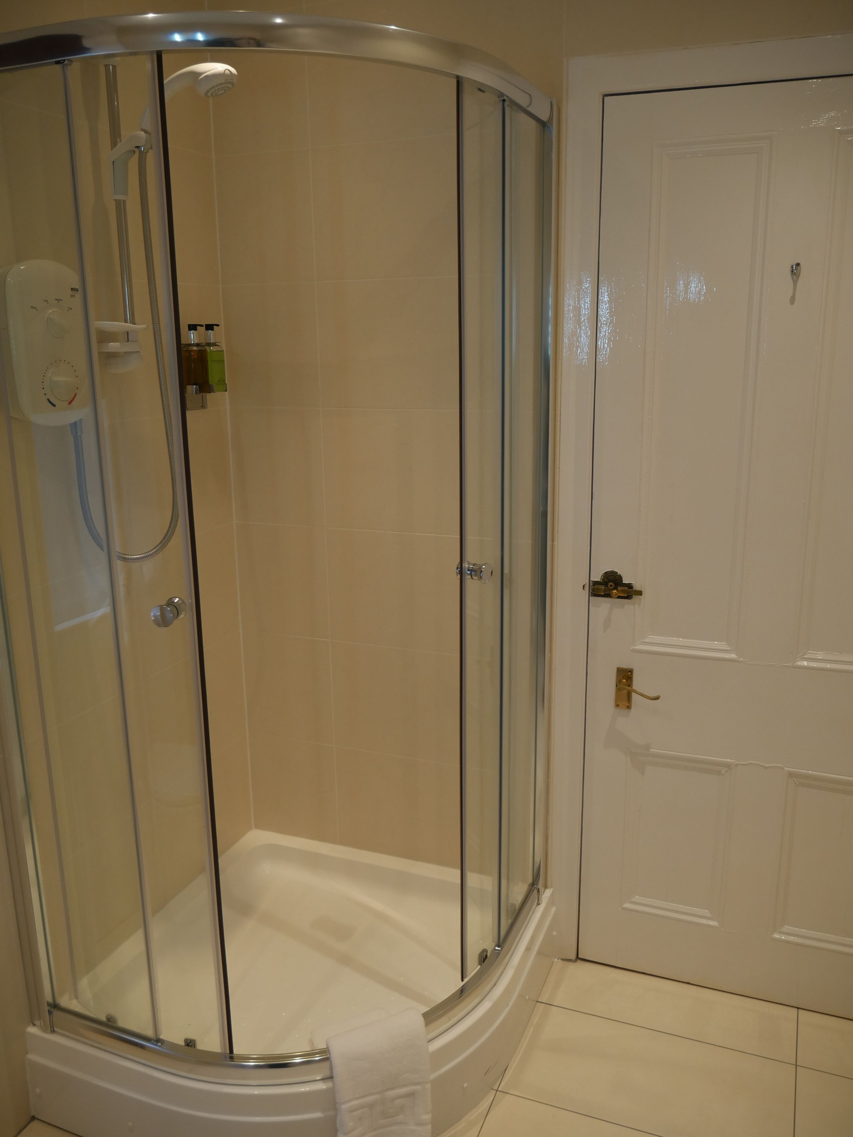 Une douche fermée par des parois de verre, dotée d’un panneau de commande, une porte blanche avec une poignée en laiton et des murs recouverts de carreaux beiges.