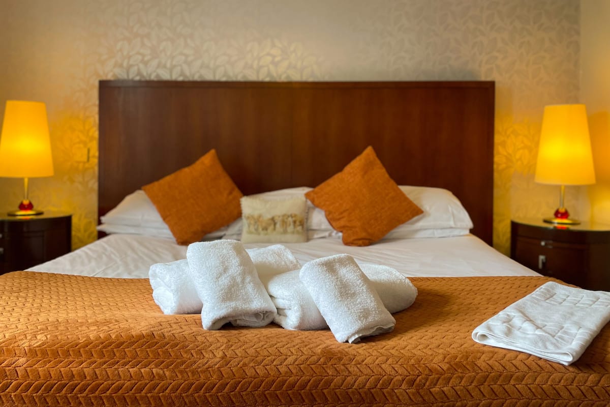 A neatly made bed with a brown quilt, two white towels, and two orange pillows. There are two bedside lamps with yellow shades on either side of the bed.