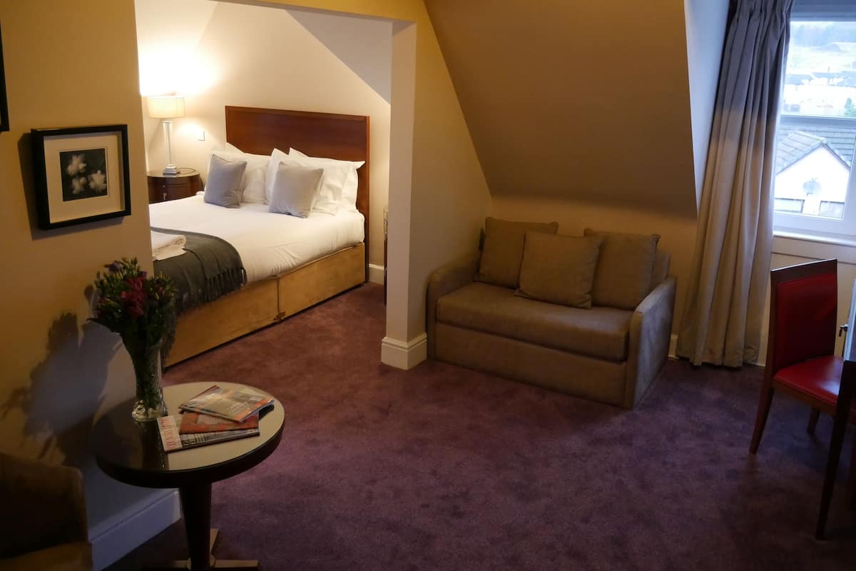 A hotel room with a bed, a sofa, a small table with a vase and magazines, and a window with curtains.
