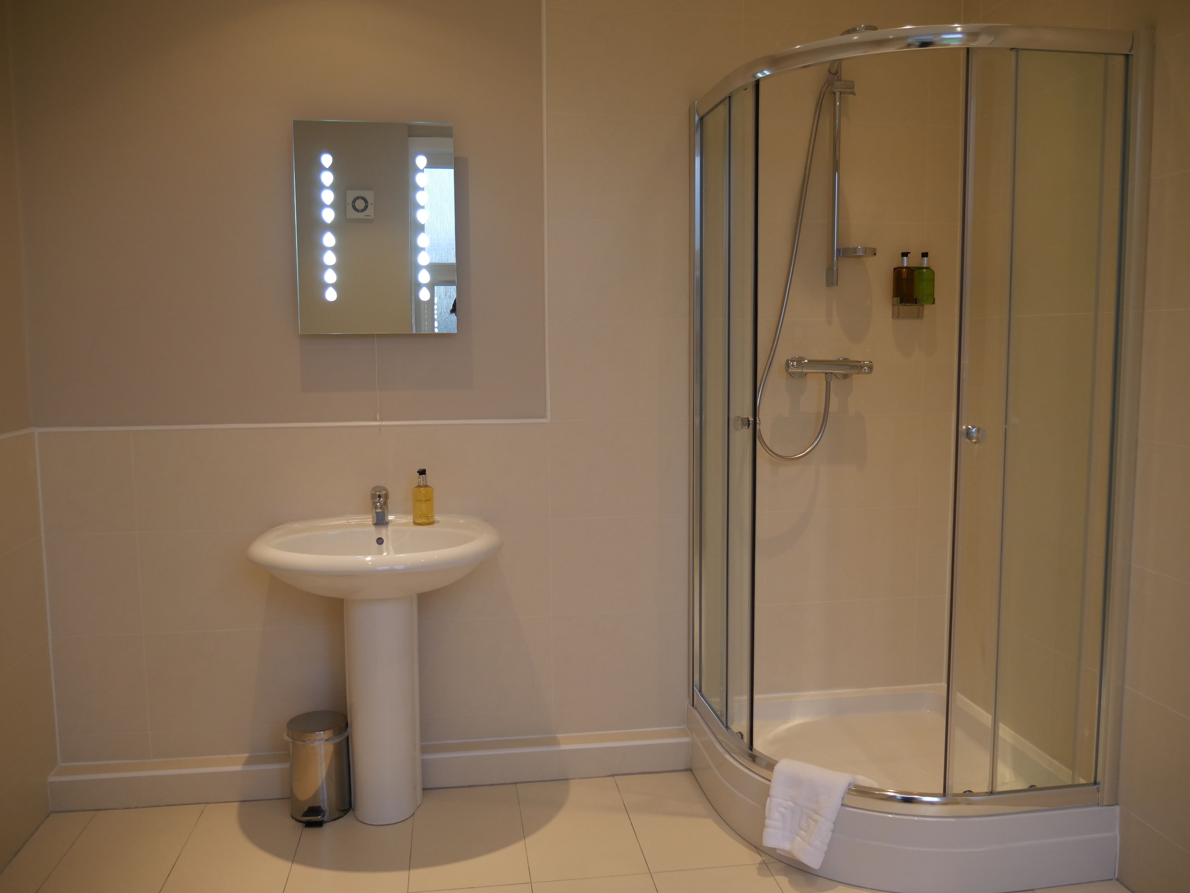 A modern bathroom with a glass-enclosed shower, a pedestal sink, and a mirror with lights.