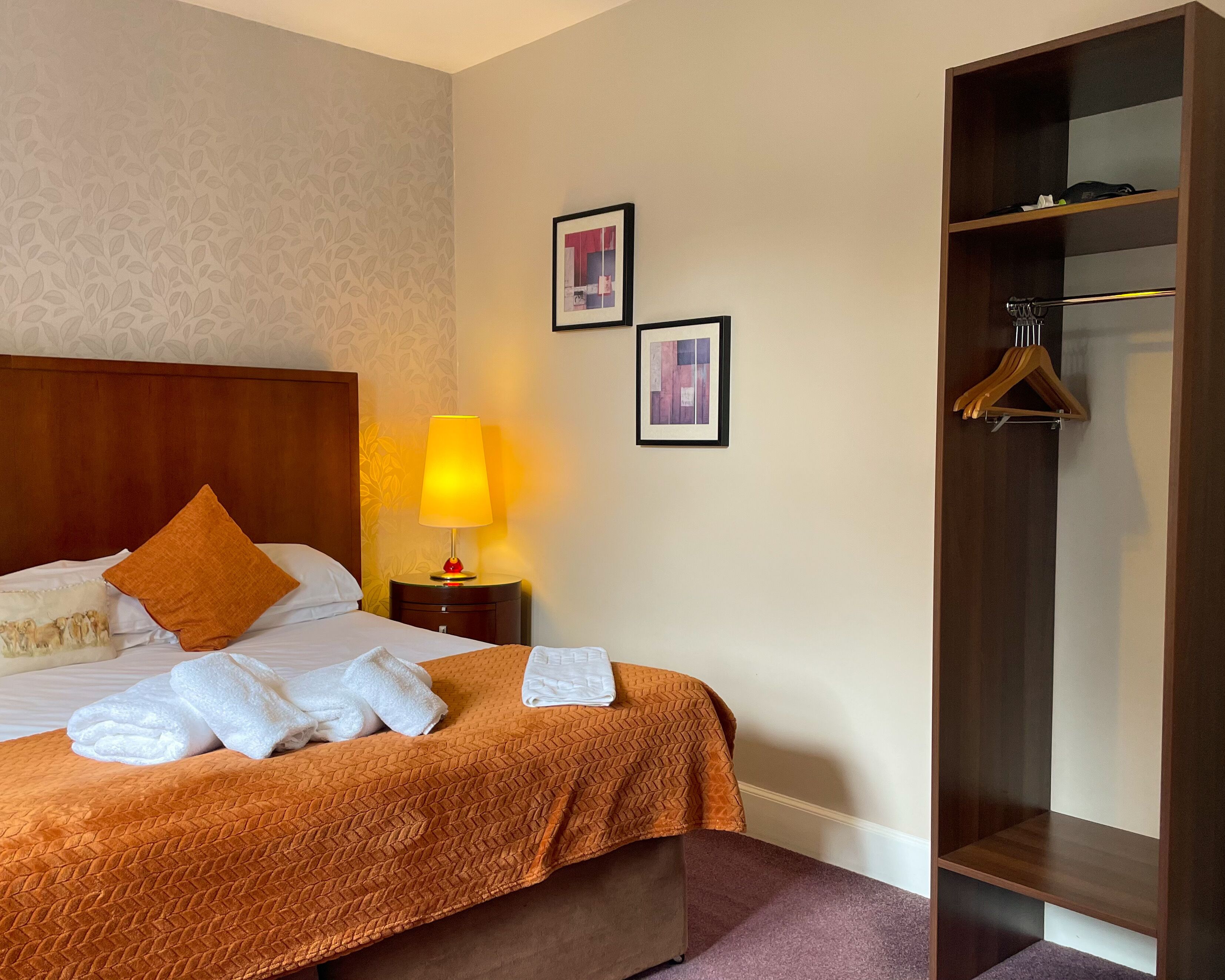Une chambre d’hôtel avec un lit, une table de chevet avec une lampe, une armoire et deux tableaux encadrés au mur.