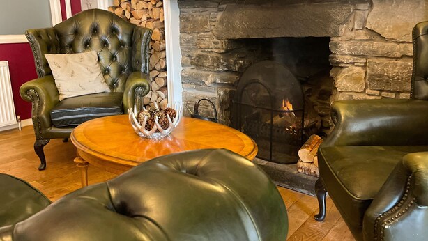 A cozy living room with leather armchairs, a round wooden coffee table, and a stone fireplace with a fire.