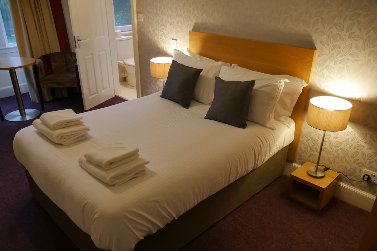 A neatly made bed with folded towels, a wooden headboard, two bedside lamps, and a patterned wallpaper.
