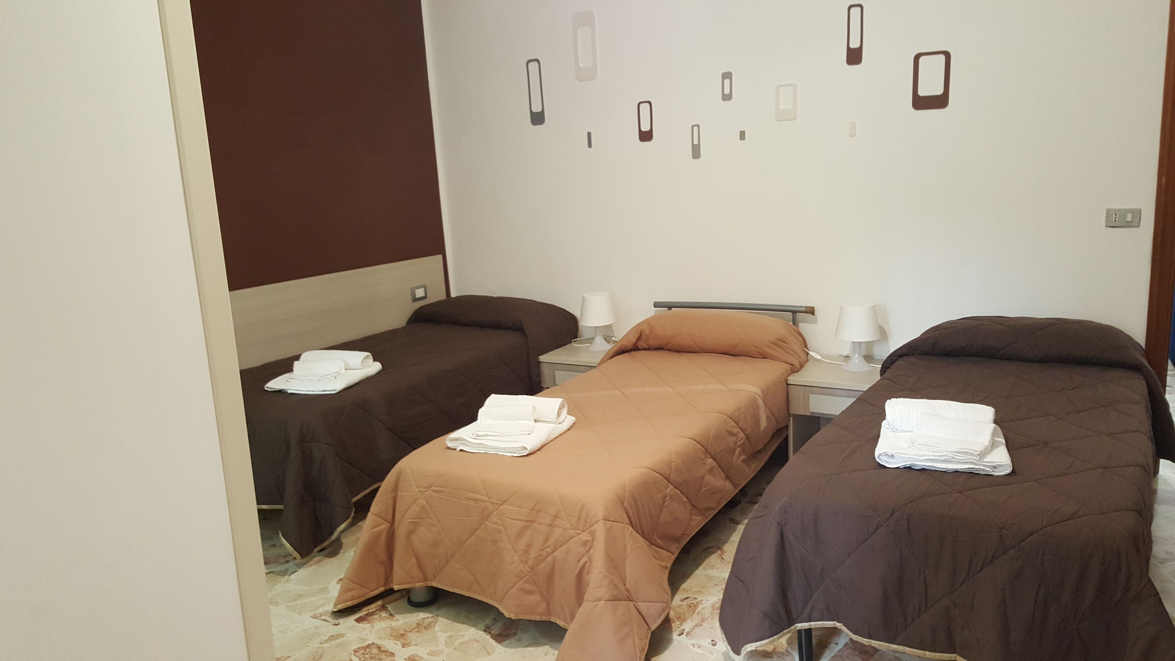 A hotel room with two single beds, each with a brown bedspread and white towels. There are bedside tables with lamps and a wall-mounted light switch.