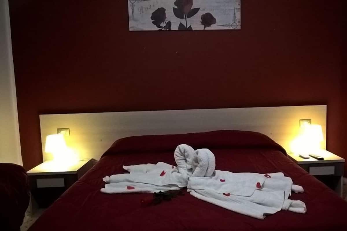 A neatly made bed with a red bedspread and white towels arranged in a heart shape. There are two bedside lamps on either side of the bed.