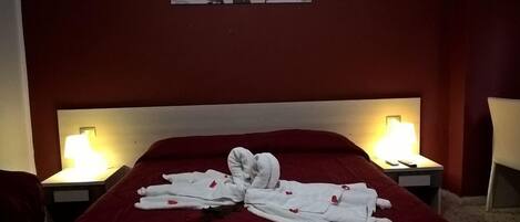 A neatly made bed with a red bedspread and white towels arranged in a heart shape. There are two bedside lamps on either side of the bed.